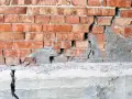Uszkodzona ceglana ściana z widocznym pęknięciem i odpadającym tynkiem, sugerującym podbijanie fundamentów.