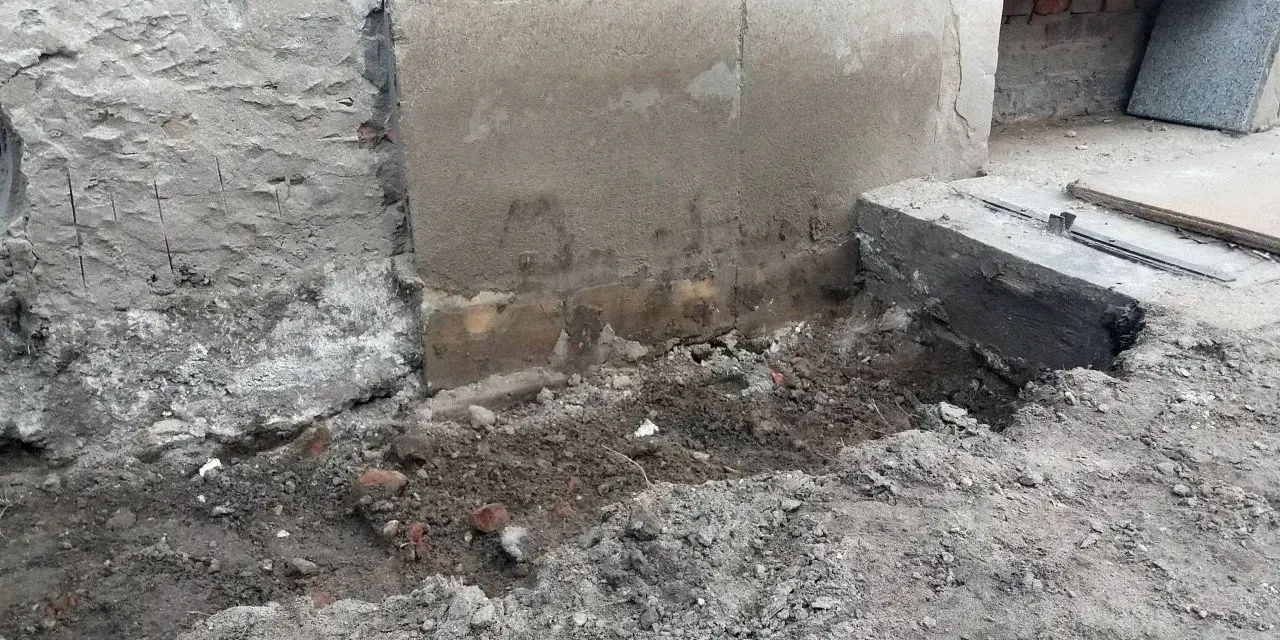 Prace przy wzmocnieniu fundamentów w starym domu. Widoczne wykopy, odsłonięta ściana i nowe elementy betonowe.