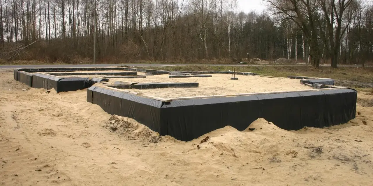 Fundament domu w piasku, widoczne czarne membrany izolacyjne i drewniane elementy szalunku. Warstwy gruntu przygotowane do dalszych prac budowlanych.