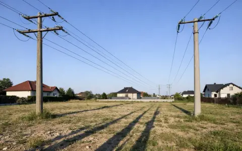 Linia średniego napięcia na działce, z której rozciągają się cienie na trawiastym terenie. W tle widać domy mieszkalne.