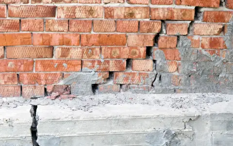 Uszkodzona ceglana ściana z widocznym pęknięciem i odpadającym tynkiem, sugerującym podbijanie fundamentów.