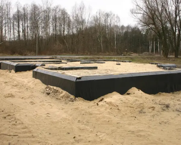 Fundament domu w piasku, widoczne czarne membrany izolacyjne i drewniane elementy szalunku. Warstwy gruntu przygotowane do dalszych prac budowlanych.