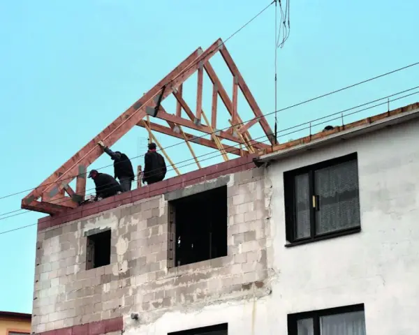 Trzech budowlańców pracuje przy podnoszeniu drewnianej konstrukcji dachu. Widać postęp prac budowlanych.