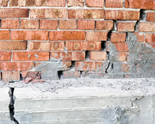 Uszkodzona ceglana ściana z widocznym pęknięciem i odpadającym tynkiem, sugerującym podbijanie fundamentów.