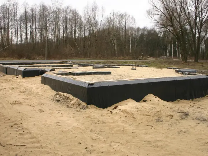 Fundament domu w piasku, widoczne czarne membrany izolacyjne i drewniane elementy szalunku. Warstwy gruntu przygotowane do dalszych prac budowlanych.