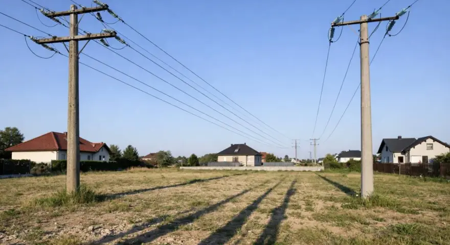 Linia średniego napięcia na działce, z której rozciągają się cienie na trawiastym terenie. W tle widać domy mieszkalne.