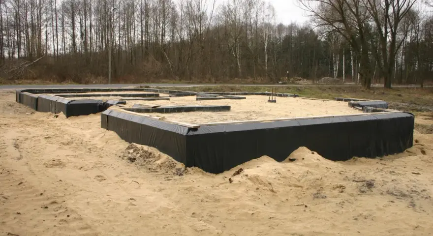 Fundament domu w piasku, widoczne czarne membrany izolacyjne i drewniane elementy szalunku. Warstwy gruntu przygotowane do dalszych prac budowlanych.