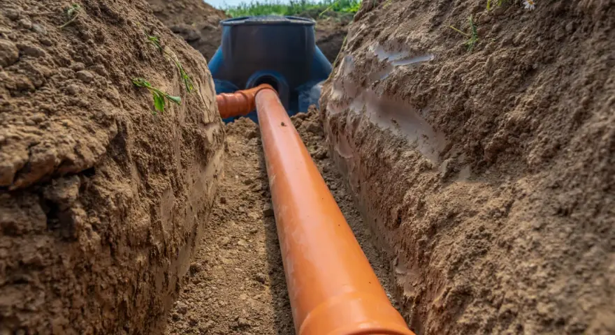 Pomarańczowa rura kanalizacyjna prowadzi do czarnego zbiornika. Trwa podłączenie do istniejącego przyłącza w wykopie.