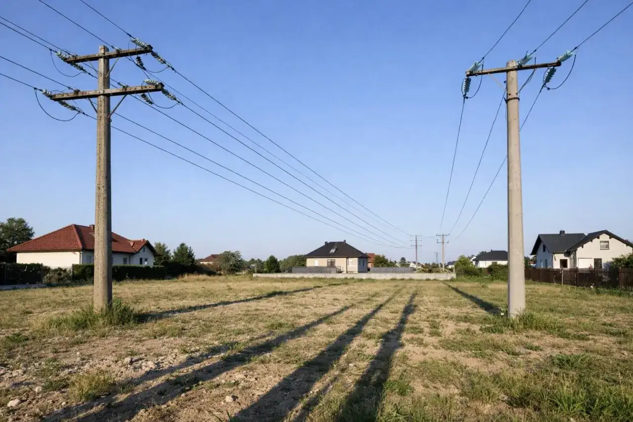 Linia 750kV biegnie nad polami i domami. Dwa betonowe słupy podtrzymują przewody wysokiego napięcia.