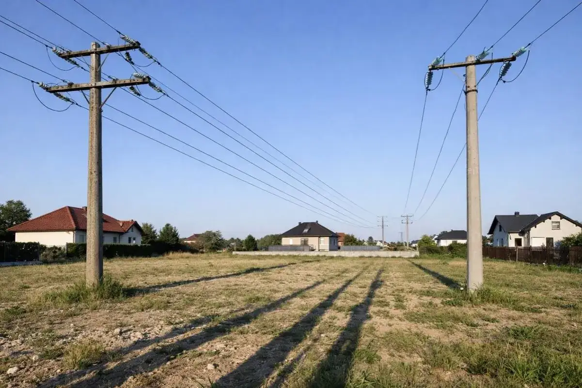 Dwa betonowe słupy podtrzymują linie przesyłowe nad pustą działką z kilkoma domami w tle.