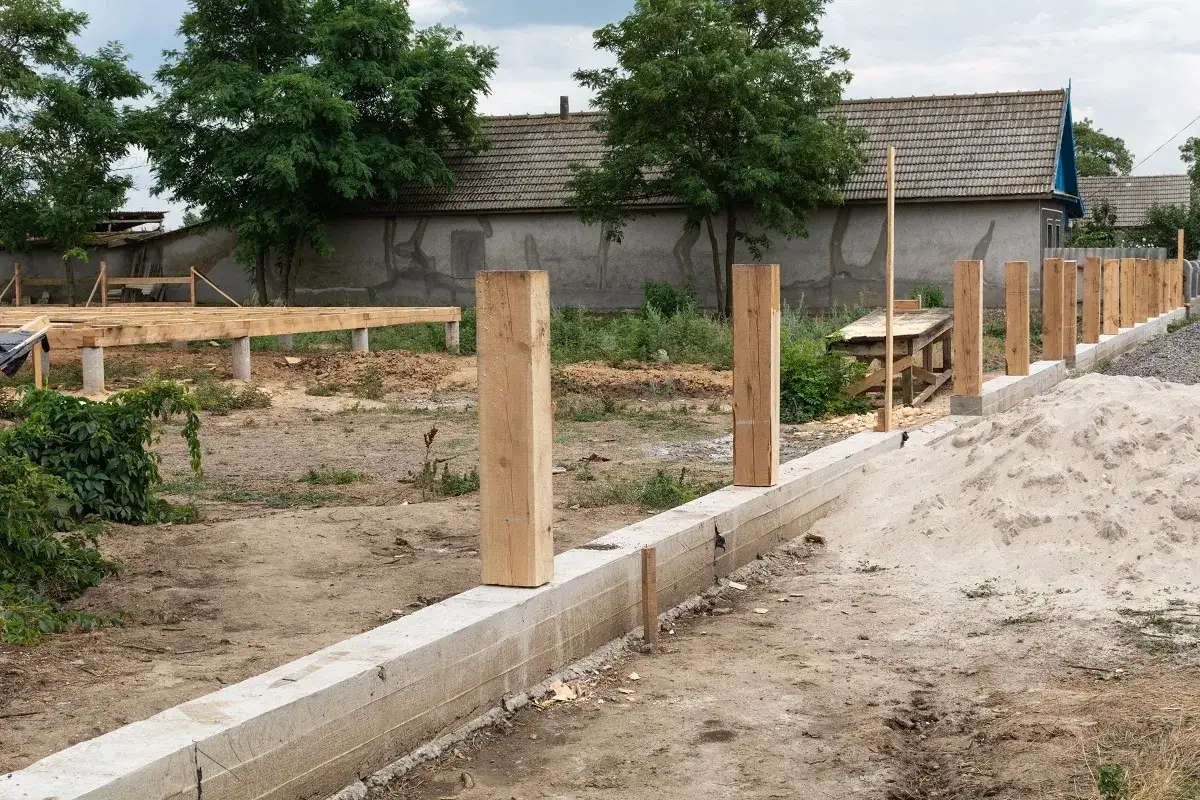 Sąsiad postawił płot na mojej działce. Widać fundament, drewniane słupki i budowę domu w tle.