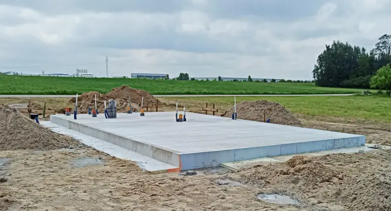 Solidna płyta fundamentowa, gotowa na budowę. Wokół piasek i zielone pola.